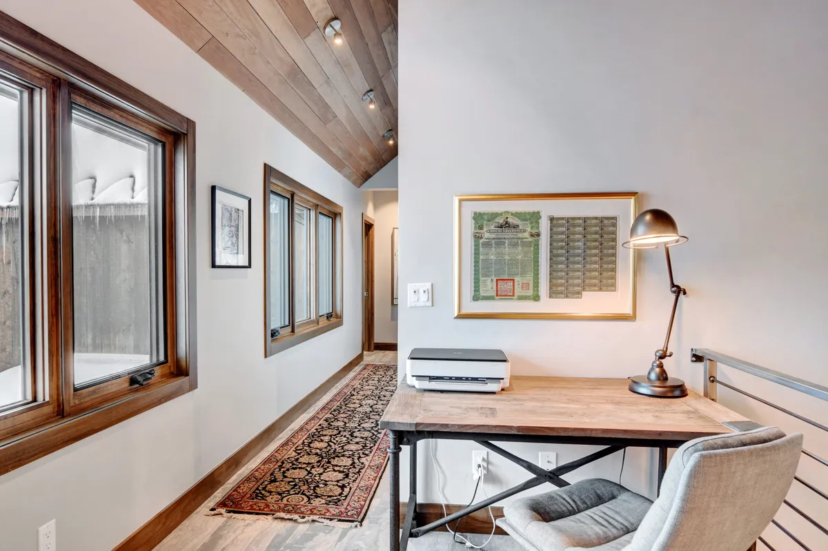 An inviting home office setup with a desk and lamp positioned in front of large windows that frame breathtaking mountain views. Warm wooden ceilings and thoughtfully placed artwork create a productive yet serene workspace.