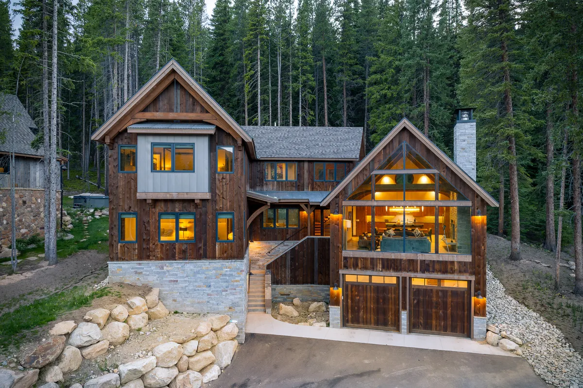 The image showcases the beautiful wooden exterior of a luxury chalet nestled among towering trees in Winter Park, Colorado. Large windows reflect the golden hues of sunset, while a stone pathway winds toward the inviting deck—perfect for enjoying mountain views, cozy fireside chats, and stargazing. The serene natural setting exudes a sense of warmth and tranquility, ideal for family gatherings or relaxing escapes.