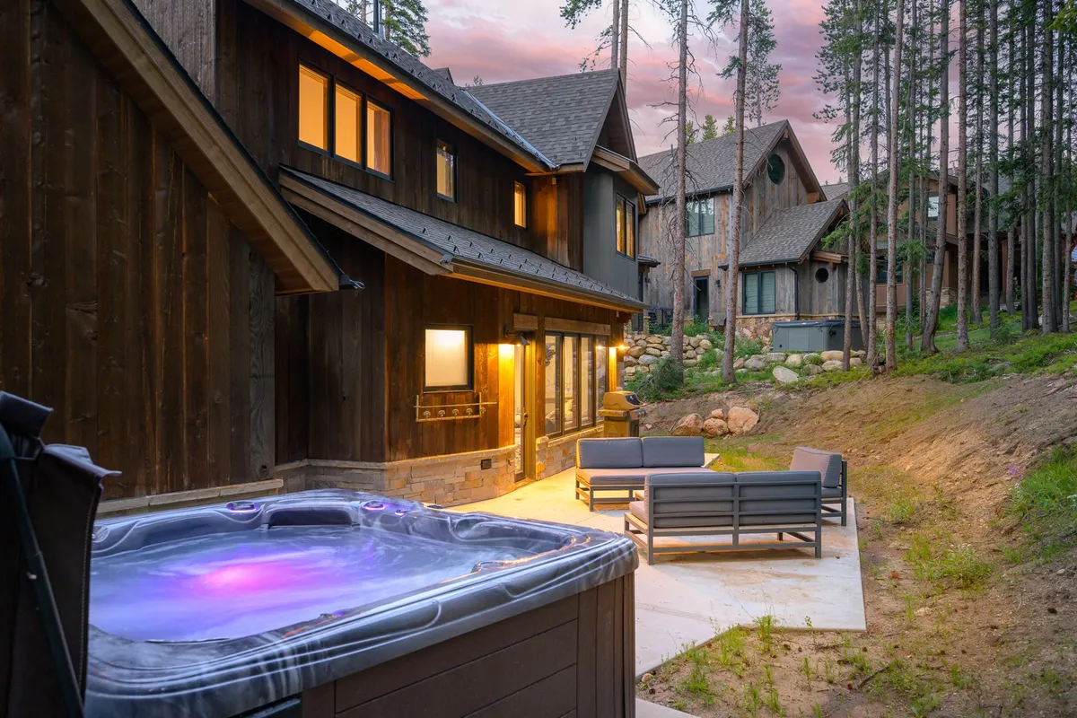 The scene transitions outdoors to an expansive deck and patio area, where a hot tub and comfortable seating invite relaxation. The wooden exterior of the chalet and surrounding natural landscape are visible, suggesting a seamless transition from inside to outside and the opportunity for year-round enjoyment.