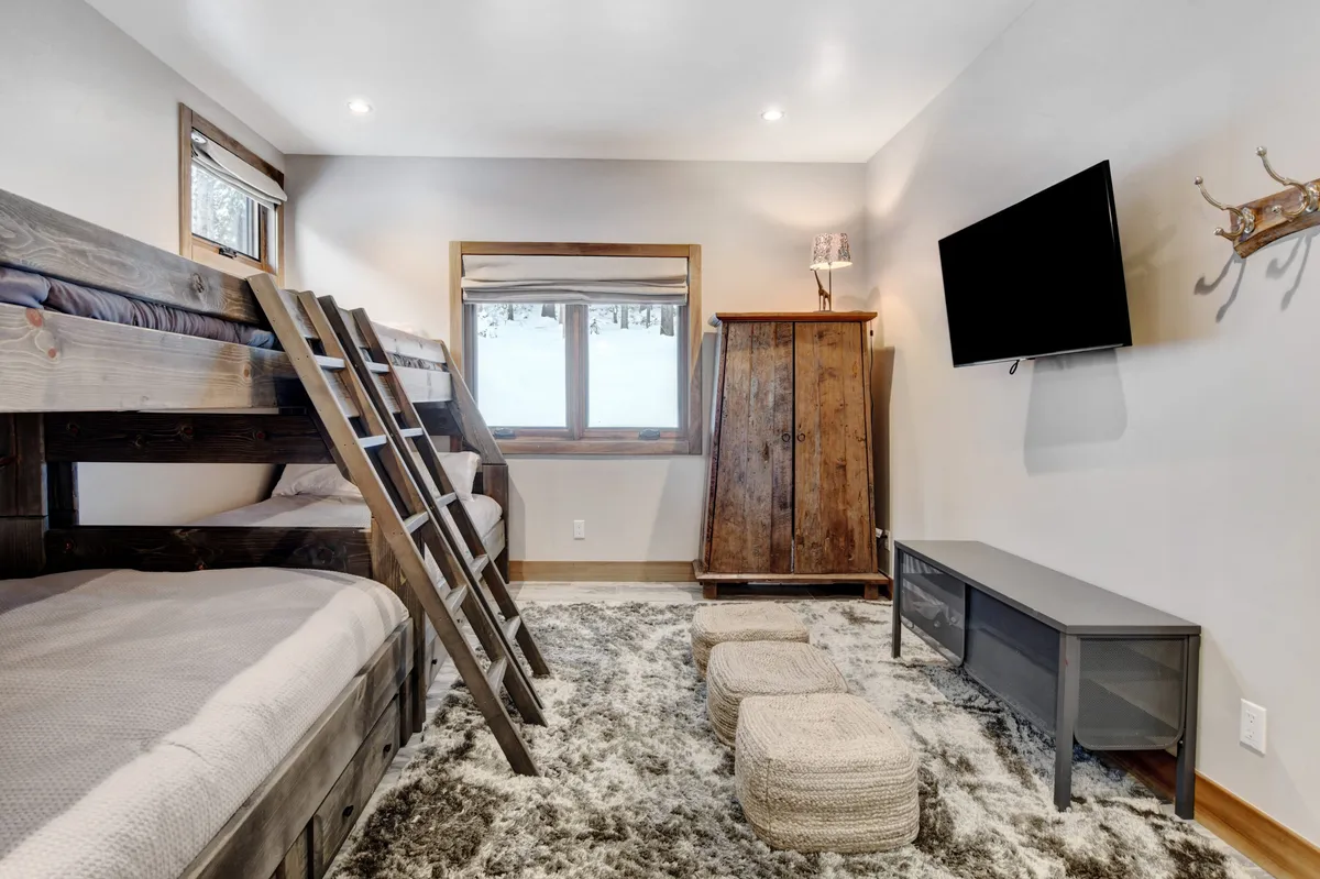 A relaxed, family-friendly room featuring bunk beds, a large window for natural light, a wooden cabinet, a wall-mounted TV, and a rug. The arrangement suggests activities like playing games, watching TV, or cozying up for late-night stories—ideal for family bonding.
