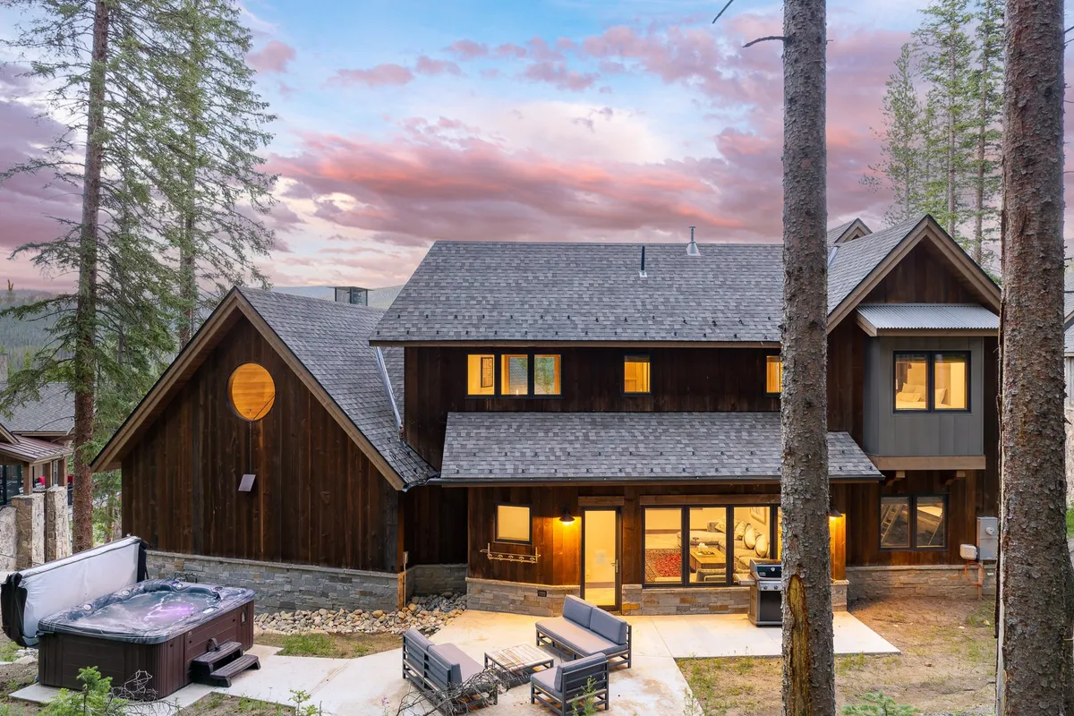 An inviting outdoor hot tub on a wooden deck, surrounded by a peaceful winter landscape with mountain views, a wooden façade, and large windows. The seating area nearby suggests a perfect spot for après-ski relaxation while soaking in the scenery.