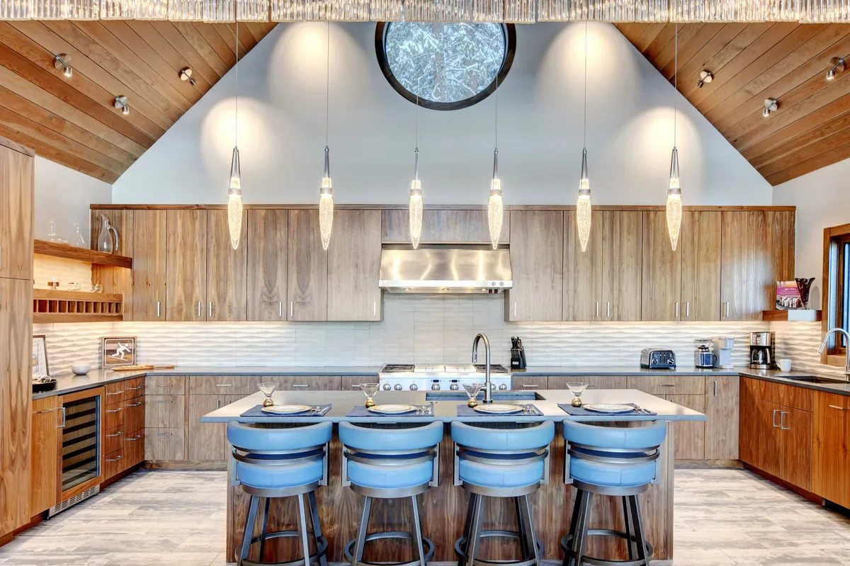 The image captures a contemporary, welcoming kitchen with a large central island, pendant lighting, and warm wooden cabinetry. A circular window adds unique character and allows natural light to brighten the space, emphasizing the area’s suitability for both cooking and gathering.
