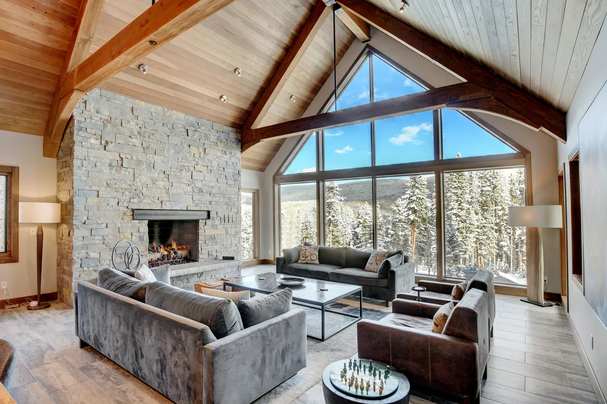 This image captures a spacious living area featuring a large stone fireplace as its focal point, surrounded by comfortable seating. Floor-to-ceiling windows reveal sweeping mountain views and fill the room with natural light. Exposed wooden beams add rustic character and warmth, creating an inviting space for gatherings.
