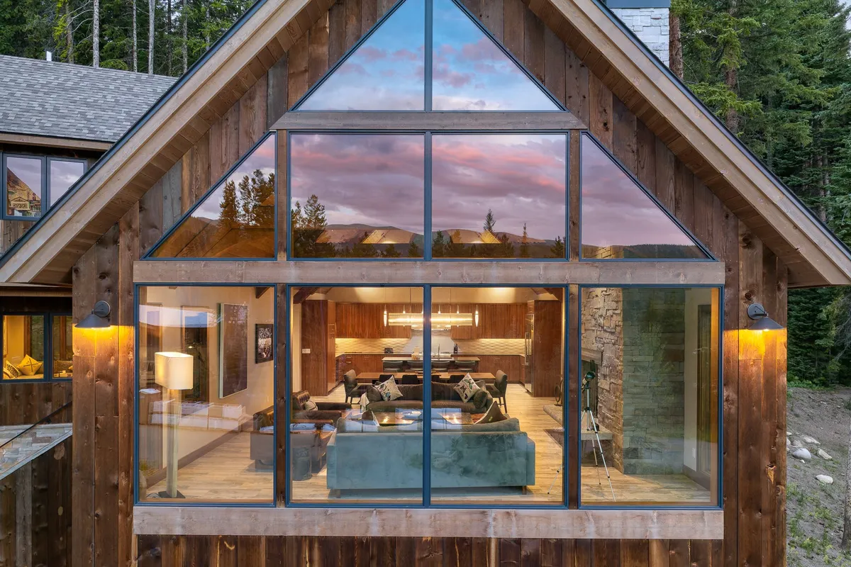 The image showcases the chalet's wooden exterior with expansive windows revealing cozy living room furniture inside. The backdrop features sweeping Rocky Mountain views, offering an immediate connection to the natural environment and capturing the retreat's immersive location.