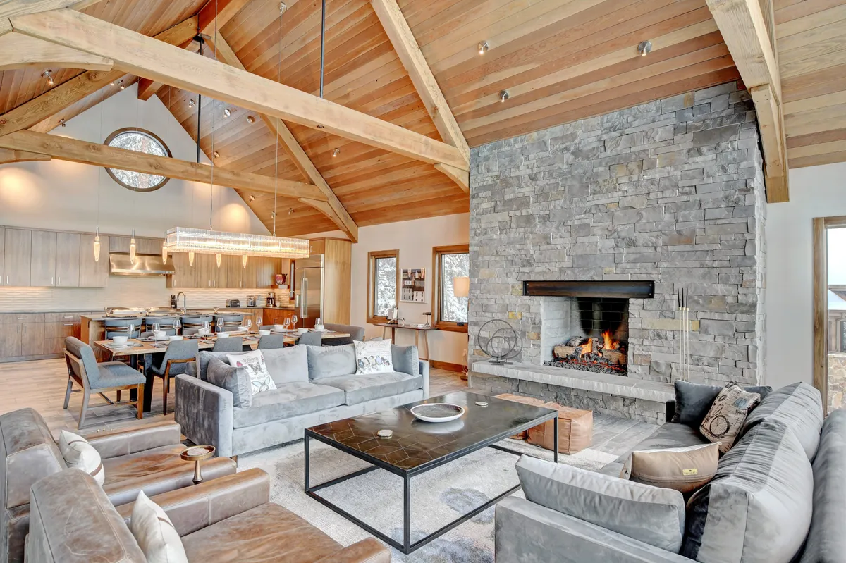 The great room is shown with a large stone fireplace, high vaulted wooden ceiling, and a spacious seating area. Expansive windows flood the room with sunlight and offer a connection to the outdoors, while a visible dining area highlights the space’s flexibility for hosting and socializing.