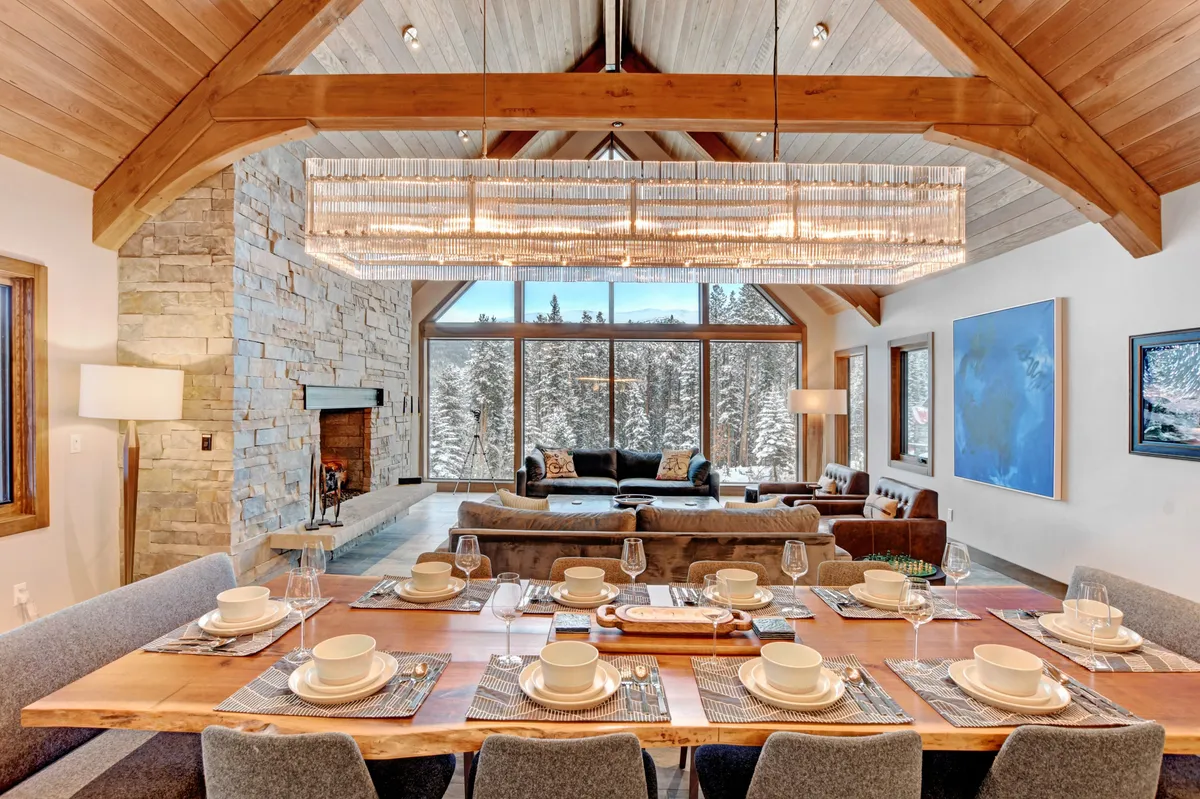 A spacious dining area with a large table set for a meal, comfortable seating for a group, and expansive windows offering snowy mountain views. A chandelier and a stone fireplace add warmth and elegance to the atmosphere.